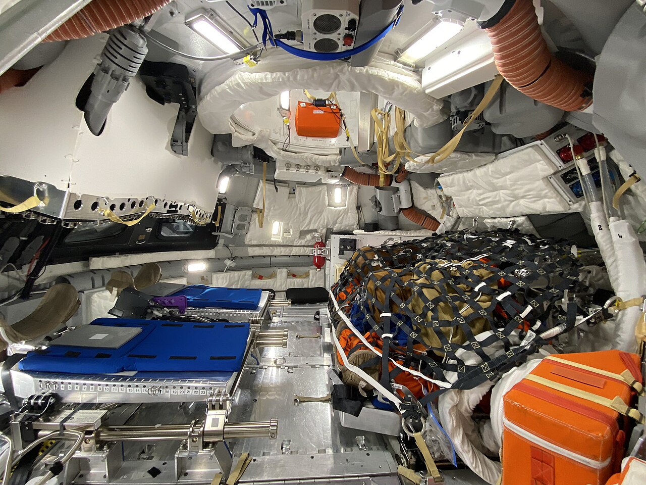 The interior of a mockup of the Orion crew module outfitted in the On-Orbit configuration that will be used in crewed missions. It features the unused Orion Crew Survival Suits on top of two of the fo